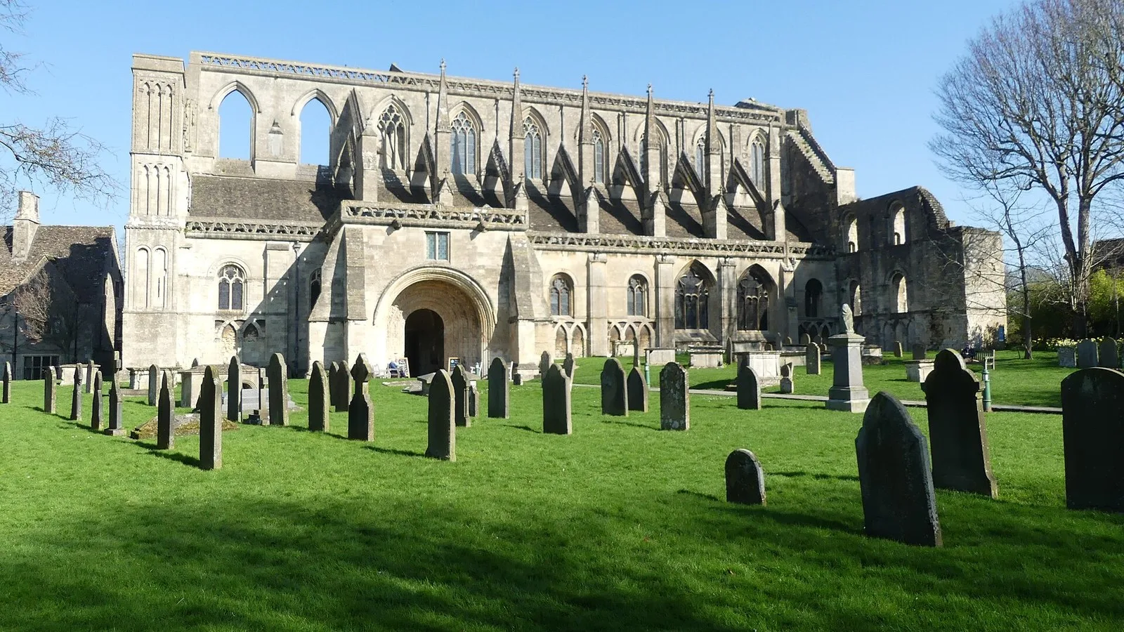 Malmesbury Abbey in Wiltshire under a clear blue sky, with gravestones on the lawn in front, the burial place of Athelstan, the first king of all England