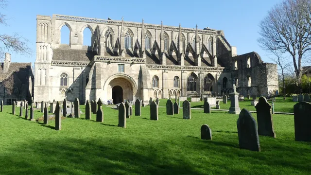 Malmesbury Abbey in Wiltshire under a clear blue sky, with gravestones on the lawn in front, the burial place of Athelstan, the first king of all England