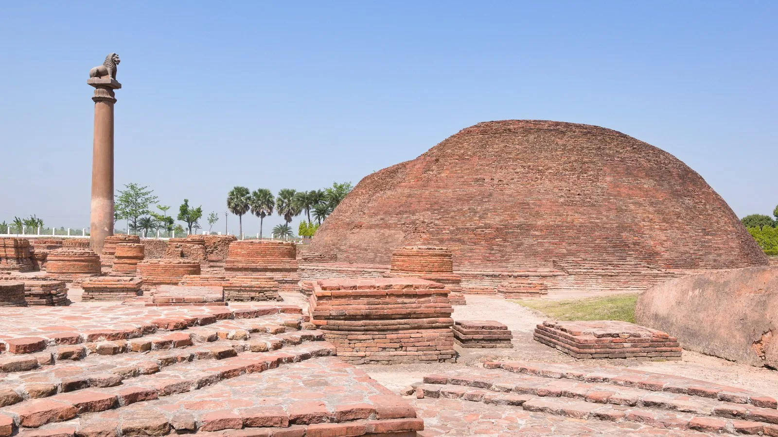 A tall stone pillar topped by a lion beside a large red-brick Buddhist stupa at Vaishali, Bihar, with low brick stupa bases in front and palm trees under a clear blue sky