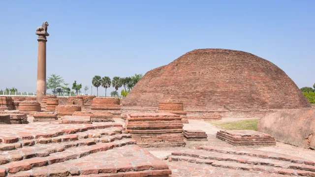 A tall stone pillar topped by a lion beside a large red-brick Buddhist stupa at Vaishali, Bihar, with low brick stupa bases in front and palm trees under a clear blue sky