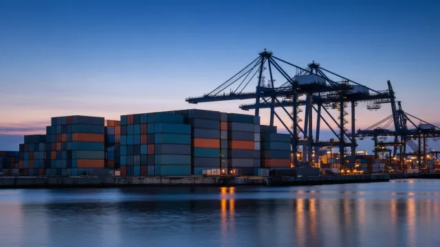 Stacks of plain teal, orange and grey shipping containers at a port at blue hour, with gantry cranes silhouetted against the sky above calm water