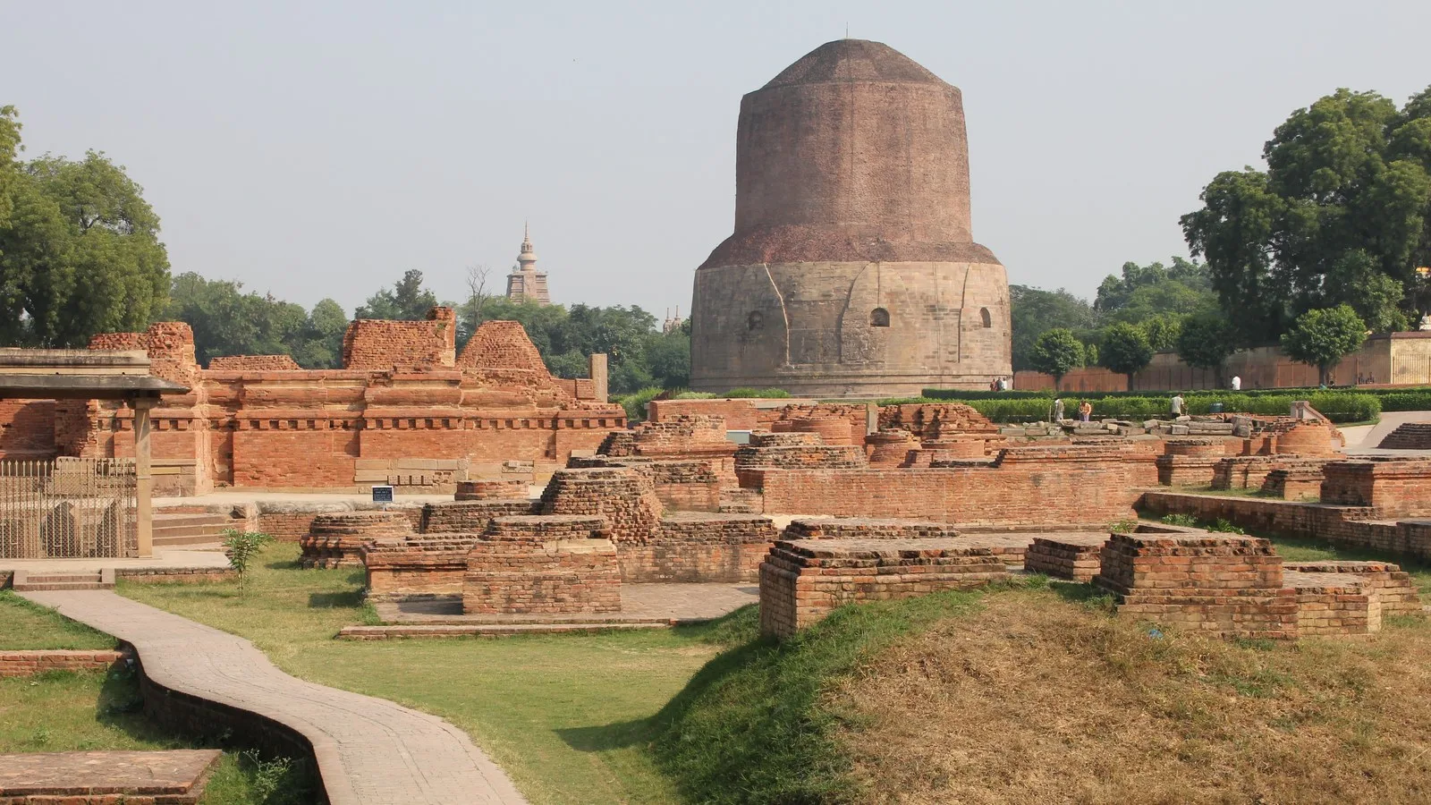 Brick ruins of ancient monasteries around the massive Dhamek Stupa at Sarnath near Varanasi, with green lawns and trees under a hazy sky