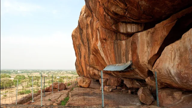Jain rock shelter at Kongar Puliyankulam near Madurai: a huge orange granite overhang with a small metal canopy over the inscribed area, a fenced rock platform and the plain below