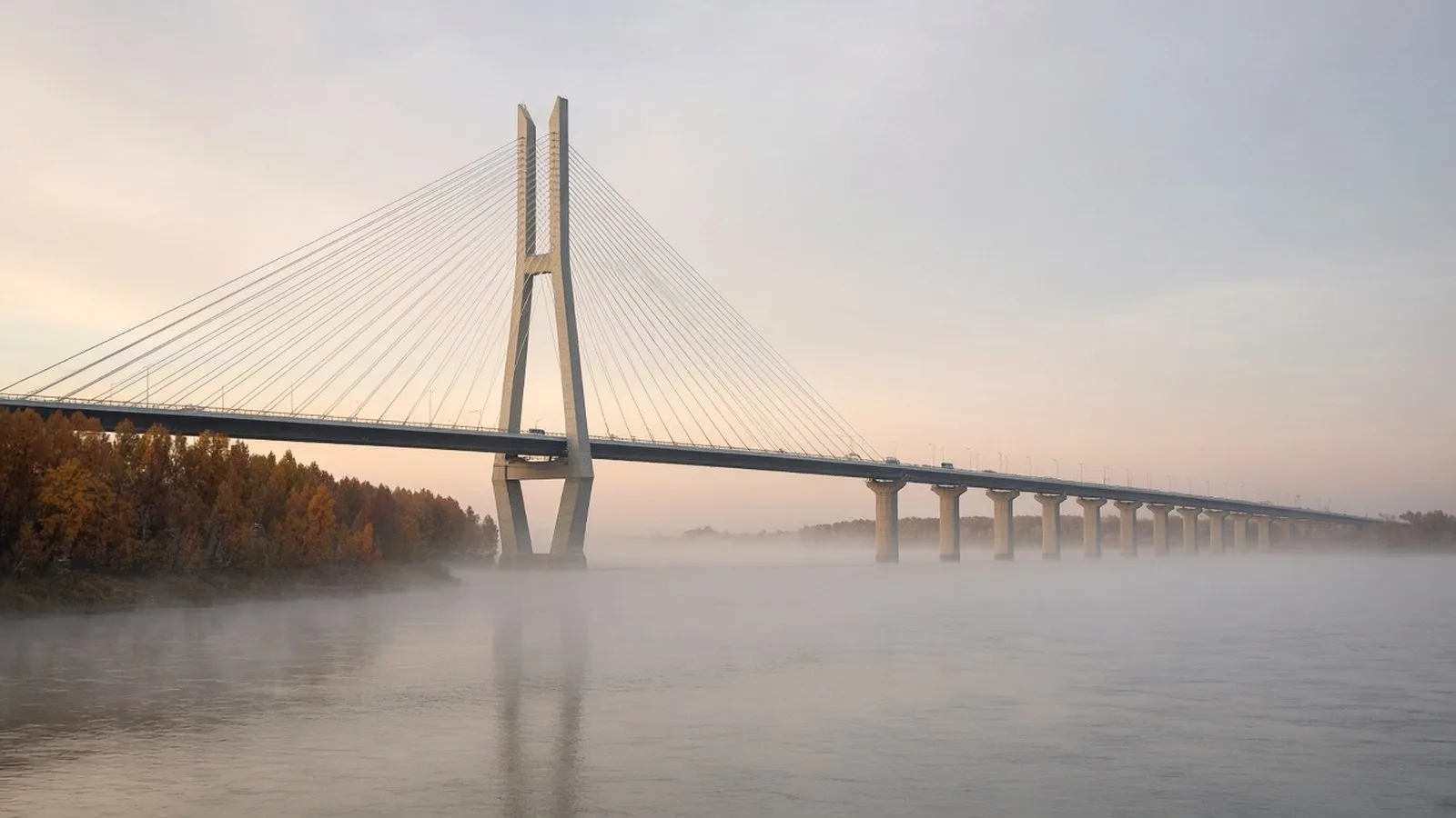 A long cable-stayed road bridge spanning a wide misty river at dawn, with autumn trees on the near bank.