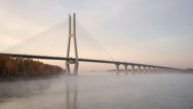 A long cable-stayed road bridge spanning a wide misty river at dawn, with autumn trees on the near bank.