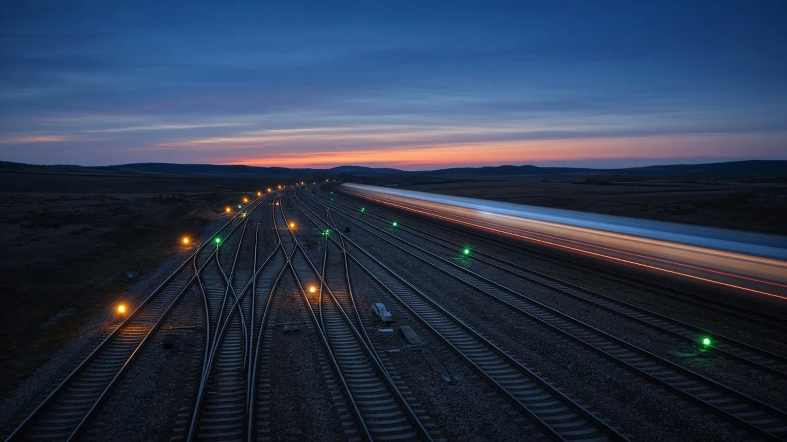 Railway junction at dusk seen from above, with tracks branching and merging, glowing green and amber signal lamps and light trails from a train