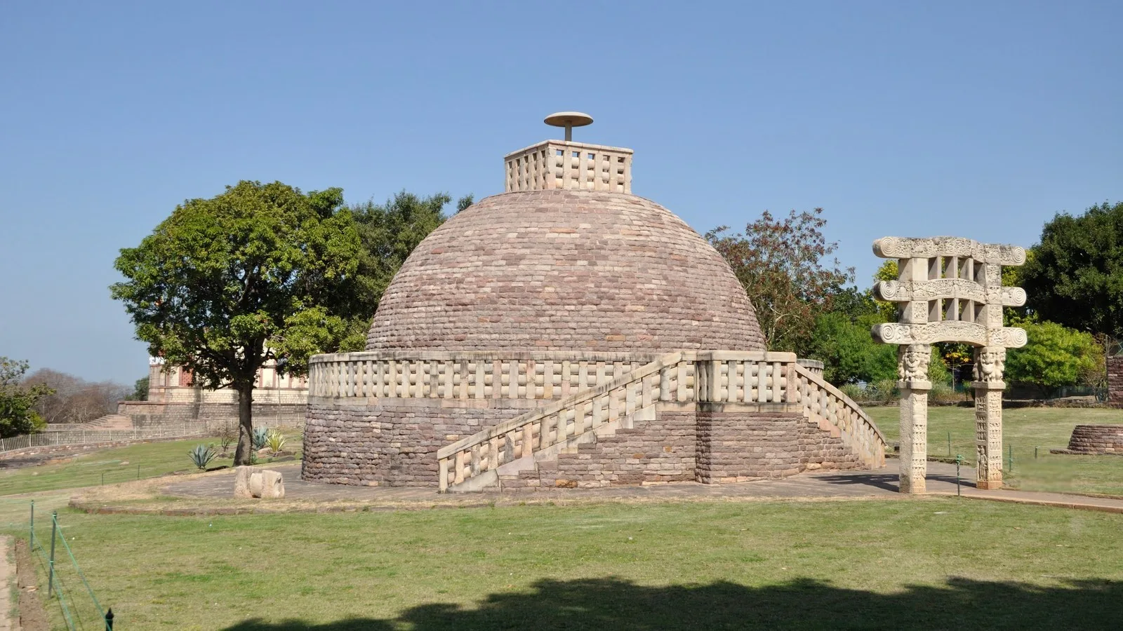 Stupa 3 at Sanchi, a stone-cased Buddhist stupa dome with a stone railing, a stairway and a single carved sandstone gateway, on a green lawn with trees under a clear blue sky