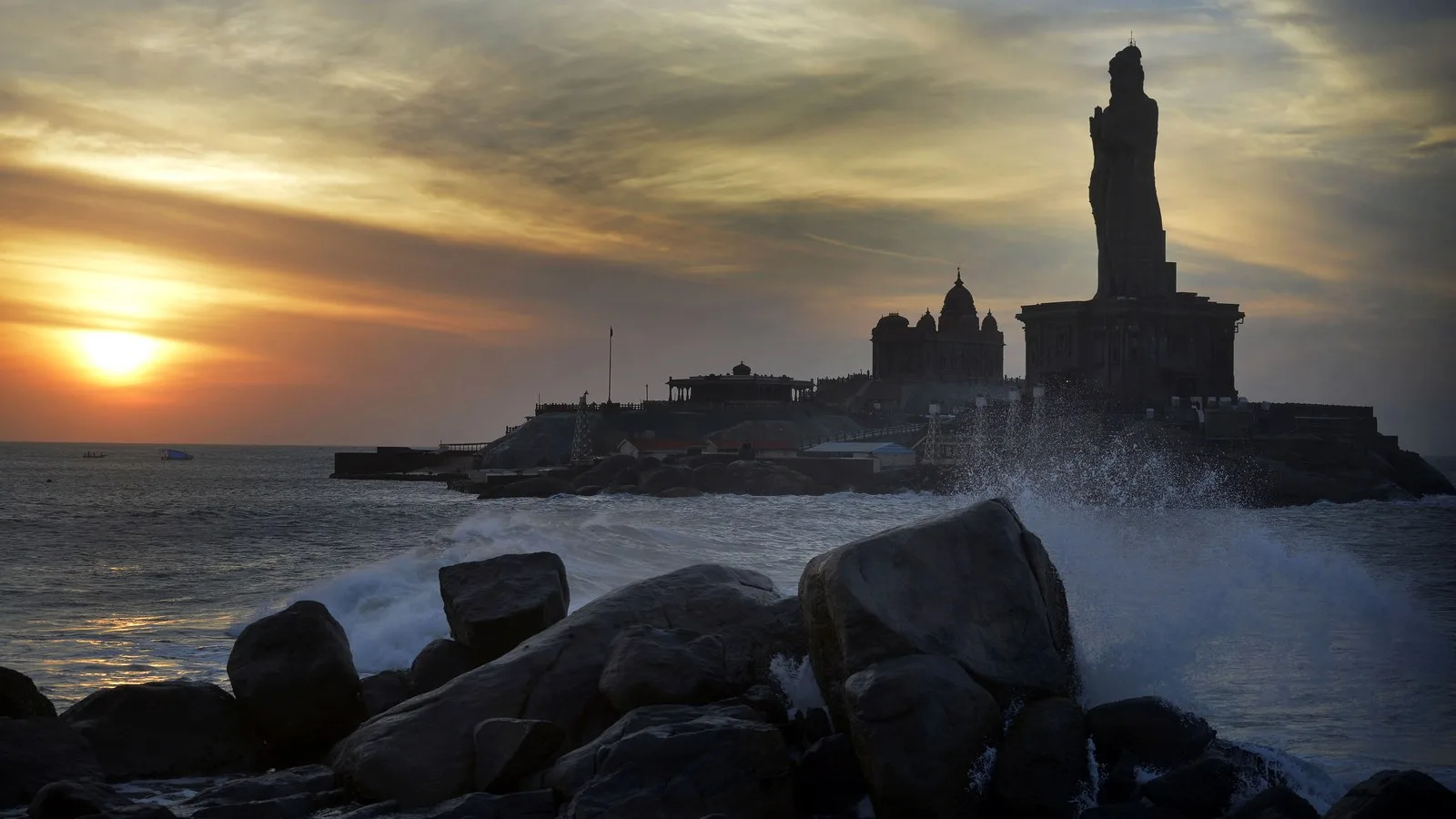 The Thiruvalluvar statue and the Vivekananda Rock Memorial silhouetted at dusk off Kanyakumari, with waves breaking on rocks in the foreground