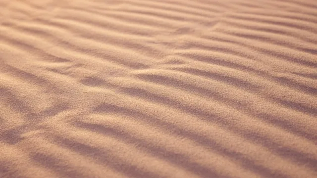 Close-up of fine sand with soft, wavy parallel ripples in warm beige and pink light