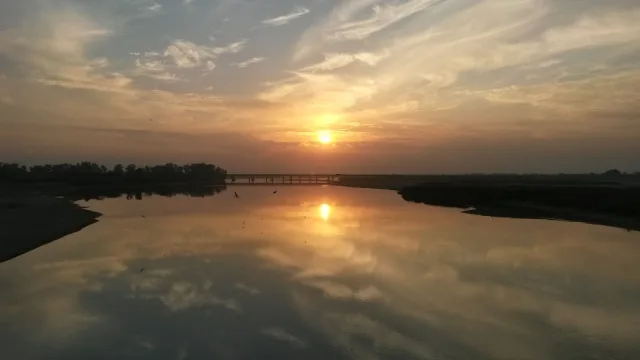 The Jhelum River, the ancient Hydaspes, at sunset near Jhelum city in Pakistan's Punjab, with the sun and clouds reflected in calm water and a low bridge on the horizon