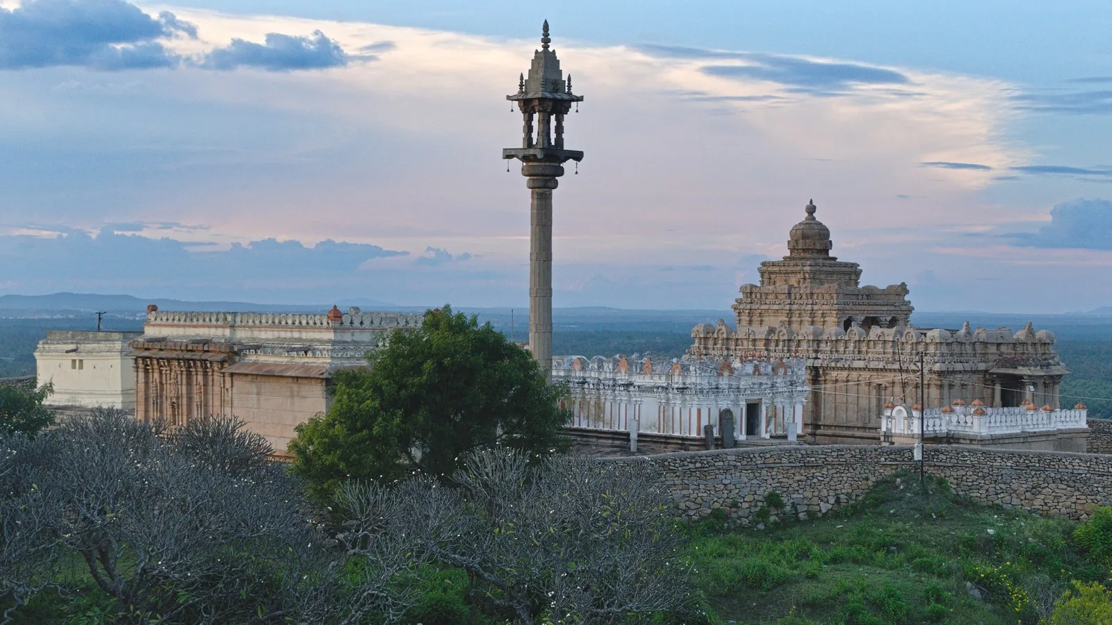 Jain temples and a tall carved stone pillar on Chandragiri hill at Shravanabelagola, Karnataka, at dusk, with trees and a stone wall in front and plains stretching to the horizon
