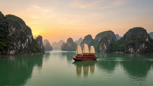 Limestone karst islands rising from calm water in Ha Long Bay with a cruise boat at sunrise